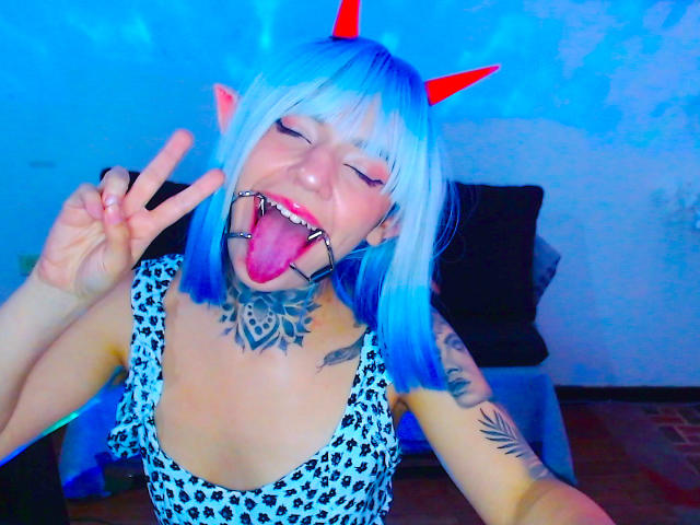Lannie Gonzales mit blauem Bob, roten Hörnern, Zunge heraus, Ballknebel, Tattoos, Leopardenkleid, zwinkernd und mit Friedenssymbol. Ihre Brüste sind klein und ihr Hintern ist nicht sichtbar..