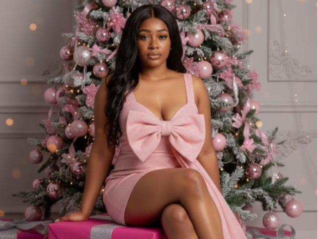 AstridDaviz sits in front of a decorated Christmas tree. She's wearing a pink dress with a large bow at the chest, showing cleavage. Her long black hair is wavy. The tree is pink and silver with ornaments. She's on a pink and silver gift. Her legs are crossed. The image has a soft, festive feel with warm, glowing lights in the background. The composition is centered and symmetrical.