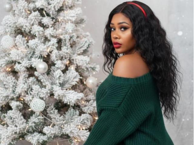 AstridDaviz with long curly black hair, red headband, green off-shoulder sweater, looking back at a snow-covered Christmas tree with white ornaments and lights.