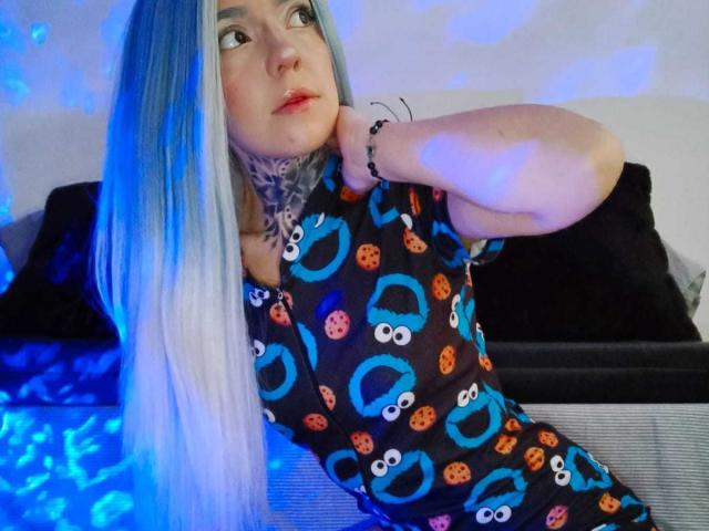 Lannie Gonzales, con lunghi capelli blu-grigi, collo tatuato e occhi azzurri, indossa una maglietta da pigiama di SpongeBob, seduta su un divano nero, con una luce blu sullo sfondo..