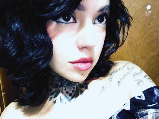 Lannie Gonzales, com cabelo cacheado preto, tatuagens no pescoço e ombro, batom rosa e delineador escuro, olhando diretamente para a câmera. Seios parcialmente visíveis, blusa com um ombro descoberto..