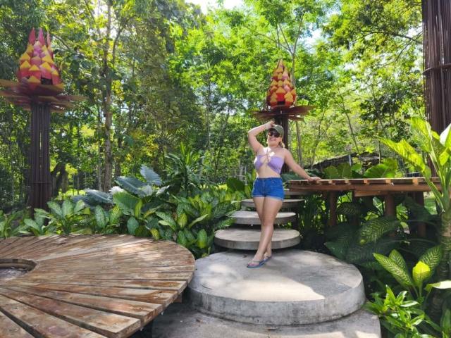 ZoeTobon69 stands in a jungle-like setting, wearing a pink top and blue shorts, her hand in her hair, her ass slightly visible, surrounded by wooden structures and greenery.