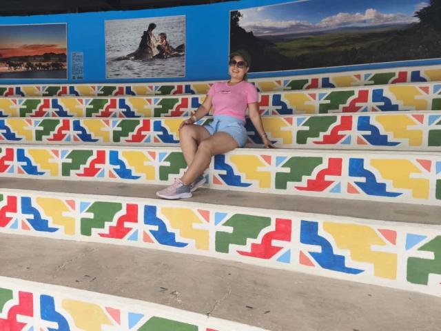 ZoeTobon69, with small tits and ass, wearing a pink tee and blue shorts, sits on colorful, patterned stairs, legs crossed, in a brightly painted, outdoor setting.