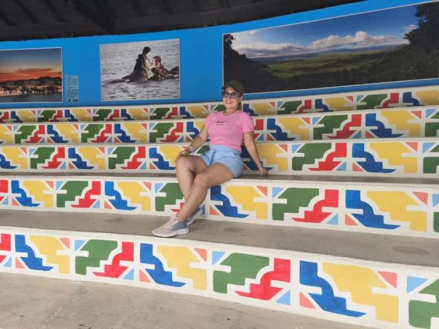 ZoeTobon69 with ass on colorful stairs, wearing pink shirt, blue shorts, white sneakers, glasses, sitting with legs crossed, smiling. Stairs have red, yellow, green, blue patterns.