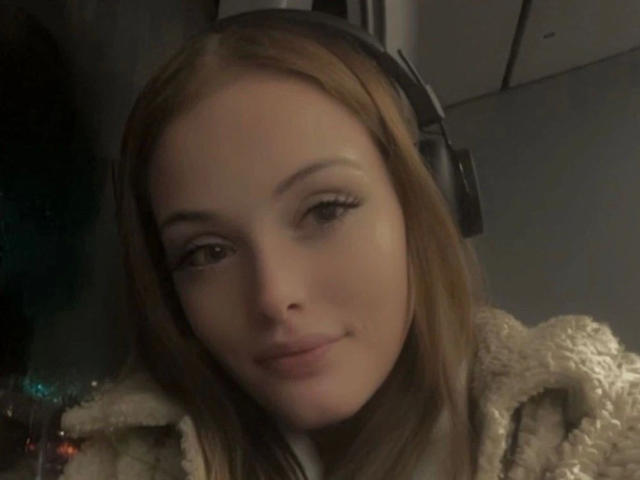 CherryTease-hot has light brown hair, wearing headphones, with a slight smile, wearing a fluffy beige coat, looking at the camera with subtle makeup.