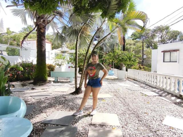 ZoeTobon69 stands in a sunlit garden, wearing a green shirt with a red graphic, blue shorts, and white sneakers, with a palm tree and white house in the background.