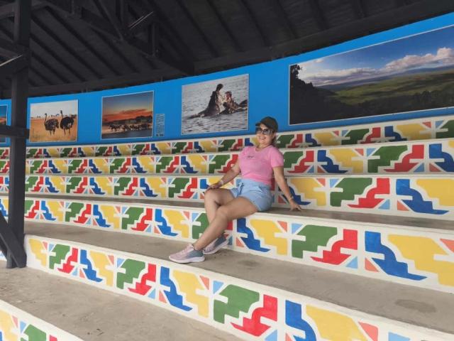 ZoeTobon69, chubby with tits in pink tee, sitting on colorful, geometric-patterned steps, wearing blue shorts and gray sneakers, under black roof with painted pictures.