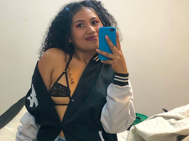 SophieSonyas with curly black hair, holding a blue phone, wearing an open black jacket, showing her small, lace-covered tits, smiling in a mirror selfie.