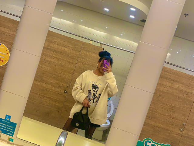 SophieSonyas with blue bun, pink phone, beige sweater, black shorts, taking mirror selfie in bathroom, reflection shows toilet in background.