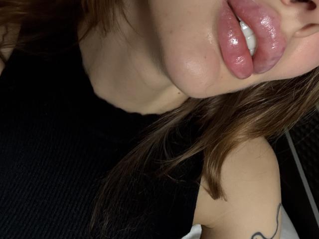 SensualMila's shiny pink lips are slightly parted, showing white teeth, with her brown hair and black sleeveless top visible in the image.