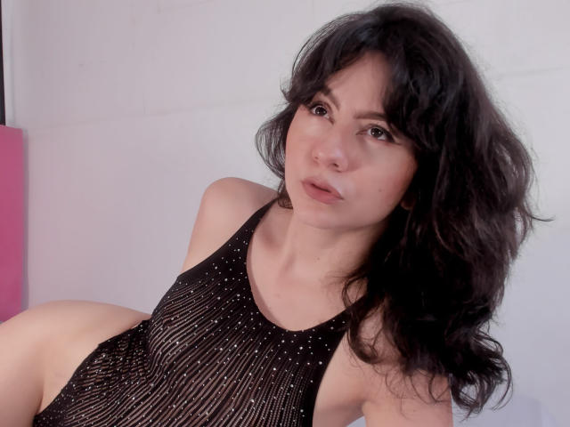 KariCarter with wavy black hair, wearing a sheer black dress, exposing her small tits, lying on her side, looking at the camera with a slightly open mouth.