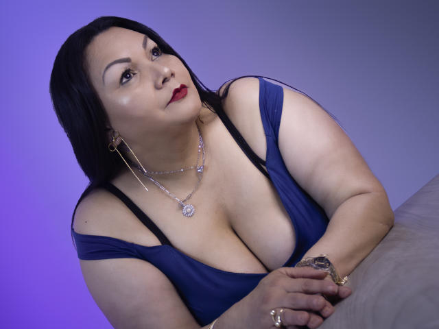 EllenaBoobs has large tits, dark hair, and red lipstick. She's wearing a blue dress with black straps, gold earrings, and a necklace. Her right hand rests on a grey surface.