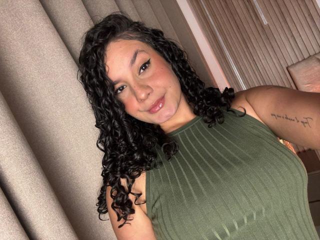 RachellBeaumont with curly black hair, green sleeveless top, and tattoo on right arm, smiling at camera, showing her medium-sized tits. Soft pink lipstick, warm lighting.