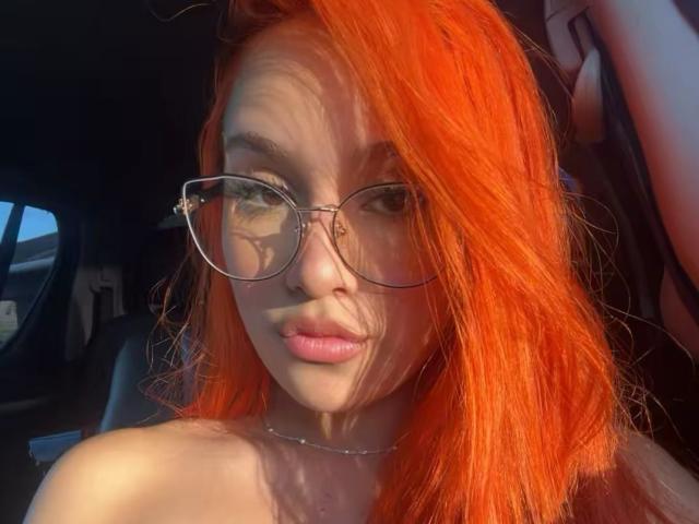 EmmaWat, avec ses cheveux orange vif, portant de grandes lunettes, a des lèvres roses et pulpeuses et un cou fin, assise dans une voiture avec la lumière du soleil sur son visage et sa poitrine..