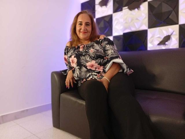 GeorgianaTaylor, with brown hair and a floral black shirt, sits on a gray leather sofa, wearing black pants, smiling with her hands on her lap, showing her ass.