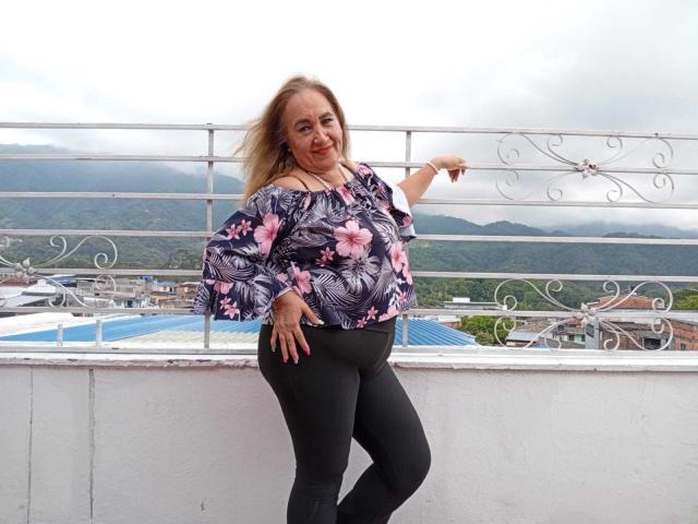 GeorgianaTaylor with blonde hair, smiling, wearing a floral off-shoulder top, black pants, standing on a balcony, hand on hip, pointing to the side, cloudy sky background.