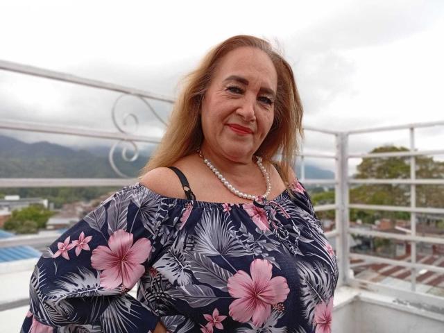 GeorgianaTaylor has medium tits, light brown hair, and wears a pink floral dress with a pearl necklace, smiling on a rooftop with white railing.