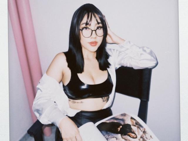 CelesteRusso with black bob, glasses, and shiny black sports bra, showing her tits, sitting with a white jacket off, and a pink curtain on the left.