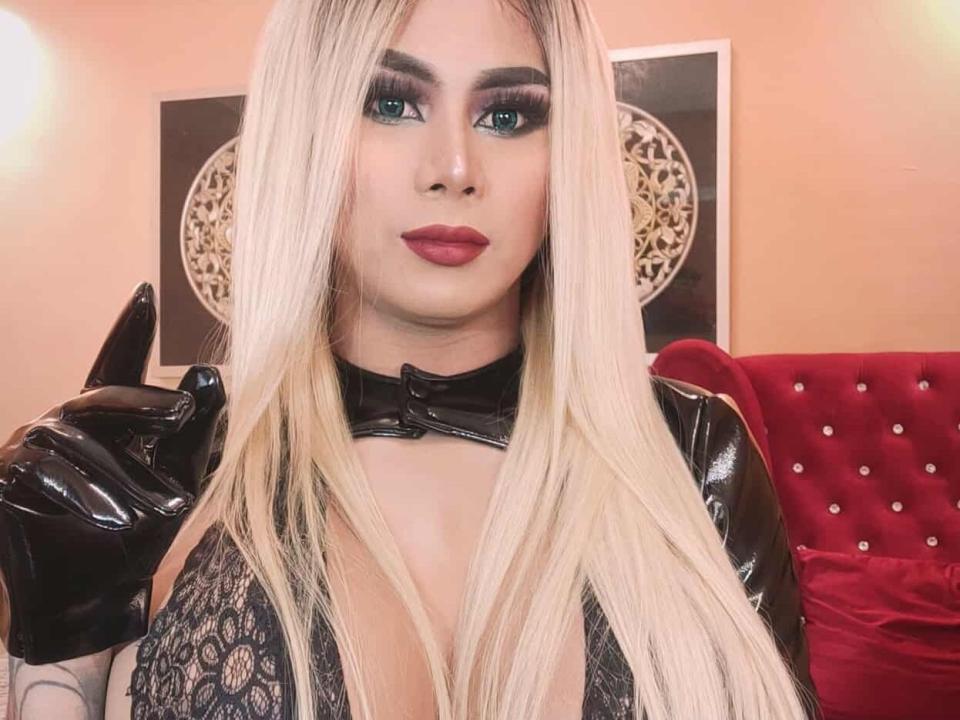 BigCockTightass com cabelo loiro comprido, olhos verdes, batom vermelho, blusa de renda preta, luvas pretas brilhantes e um colar preto, fazendo um leve gesto com o dedo, olhando diretamente para a câmera..