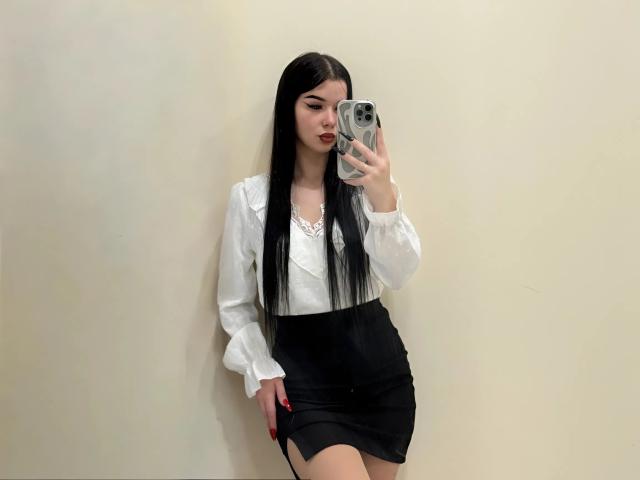 AmberMartha with long black hair, white blouse, black high-waisted skirt, red lipstick, holding phone, taking mirror selfie, showing her tits and ass, standing against plain wall.