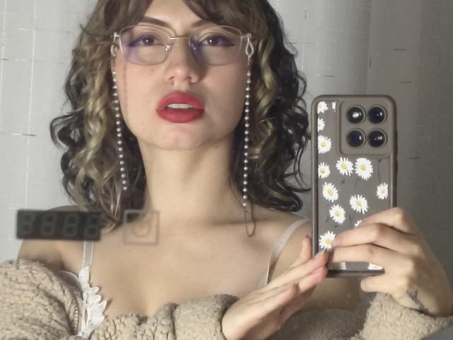 ElizaCanterville with curly brown hair, red lipstick, glasses, daisy-patterned phone case, pearl earrings, beige sweater, holding phone, showing tits, white tile wall in background.