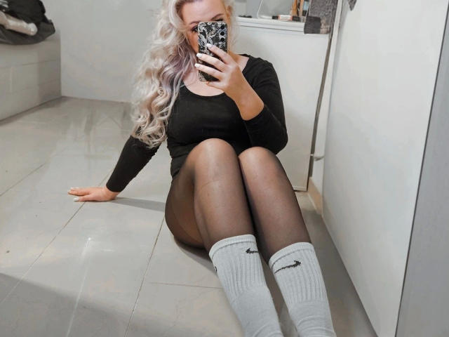 TessaWayne-hot with platinum blonde hair, black long-sleeve top, black pantyhose, and grey Nike socks, sitting on a tile floor, taking a mirror selfie with a phone.