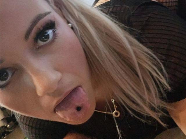 TessaWayne-hot with blonde hair, thick eyebrows, and long eyelashes, sticking out her tongue with a pierced lip, wearing a black mesh top, in a selfie.