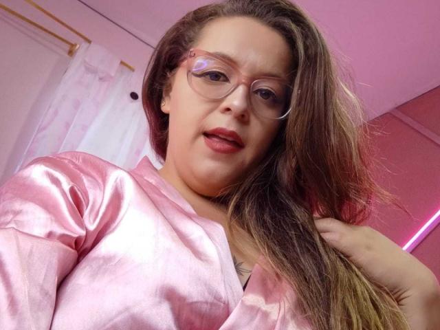 CleareRems with long brown hair, wearing pink satin shirt, glasses, and red lipstick, holding hair, pink room, slightly open mouth, soft pink lighting.
