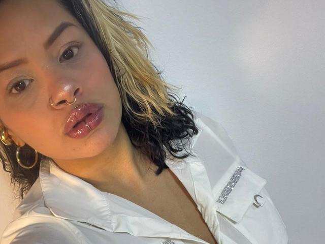 MariGonz has shiny pink lips, a nose ring, and curly black-and-blonde hair, wearing a white shirt with a sparkly button. She has golden hoop earrings and a serious look.