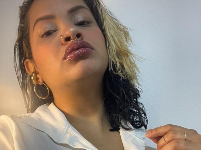 MariGonz with glossy pink lips, nose ring, and gold hoop earrings, wearing a white shirt, half-black, half-blond curly hair, holding collar, looking down, subtle shine on skin.