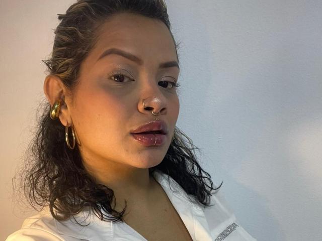 MariGonz, avec ses cheveux bruns mouillés, ses boucles d'or, son anneau de nez et ses lèvres roses brillantes, porte une chemise blanche, avec un mur uni en arrière-plan. Sa peau est bronzée et elle a l'air confiante..