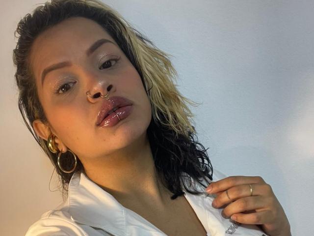 MariGonz has medium brown skin, black and blonde wet hair, gold hoop earrings, nose ring, shiny pink lips, and a gold ring, wearing a white shirt.