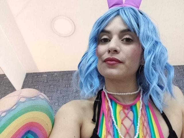 Lilaa, een meisje met blauw golvend haar, roze konijnenoren, een kleurrijke gestreepte halter, een parelketting en roze lippenstift, die haar borsten laat zien in een selfie. Op de achtergrond een regenboogkleurig kussen in de vorm van een kont..