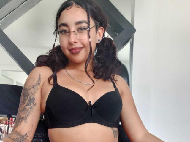 AryVixen com seios marrons volumosos em um sutiã preto, óculos e maria-chiquinhas, mostrando tatuagens, sorrindo e usando um colar. Ela está sentada em uma cadeira preta..