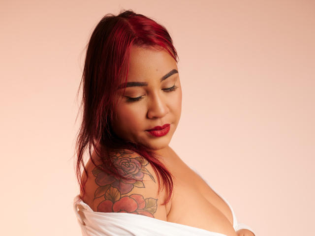 KinkyHouseWife with red hair, tattooed shoulder, and white shirt off, showing tits, red lipstick, and closed eyes, against a pink background. Her tattoo features roses and peaches.