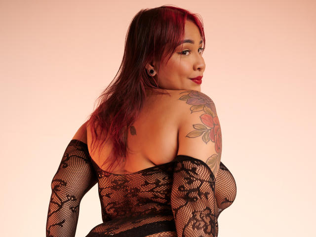 KinkyHouseWife with red-tipped hair, lace top, and tattoos, showing her back and looking over her shoulder, revealing her tits and ass through black fishnet sleeves.