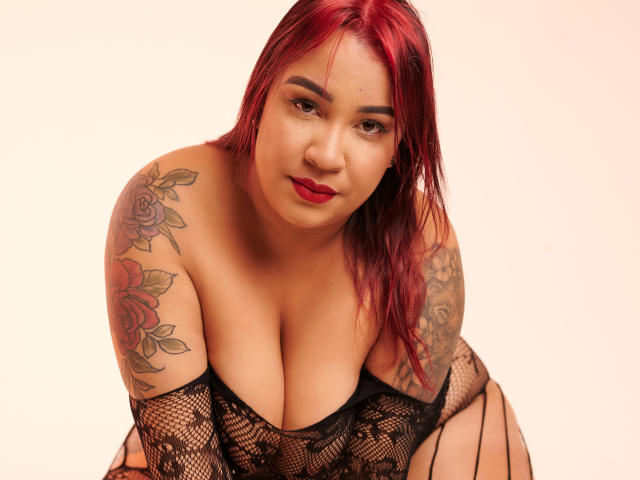 KinkyHouseWife with red hair, tattoos on shoulders, and black fishnet lingerie, showing big tits, leaning forward, red lipstick, and a sexy smile.