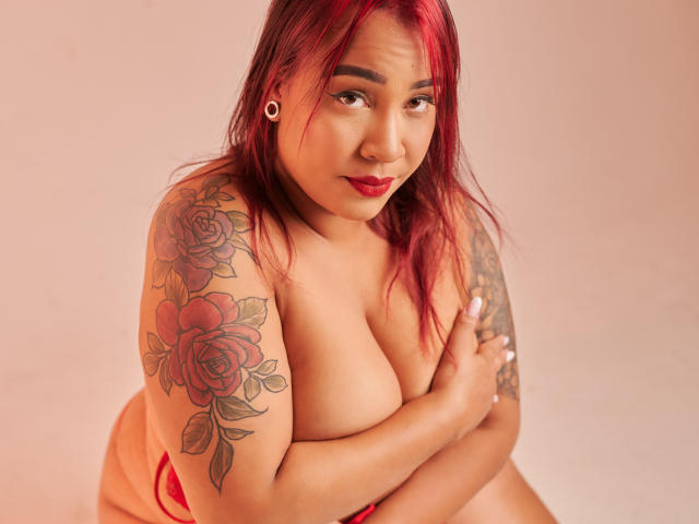A photograph of KinkyHouseWife with red-dyed hair and a curvy, plus-size figure. She has large, exposed breasts, covered by her arm, and wears a red strapless top. Her right arm has colorful tattoos of roses. She has a confident, slightly smirking expression, with red lipstick and white ear studs. The image has warm, soft lighting. Her skin is light brown, and she has a subtle, natural makeup look. The composition is close-up, focusing on her upper body and face.