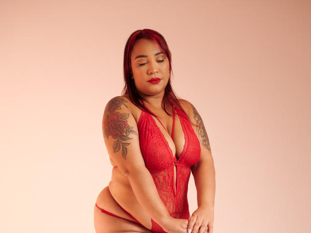 KinkyHouseWife with red hair, wearing red lace lingerie, showing tattoos on her right arm, holding her red panties, with her eyes closed and red lipstick.