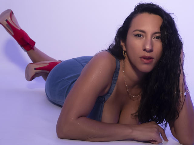JadeFoster, lying on her stomach, wearing red heels, blue denim dress, showing her tits, with wet, dark hair, gold earrings, and a necklace, looking at the camera.