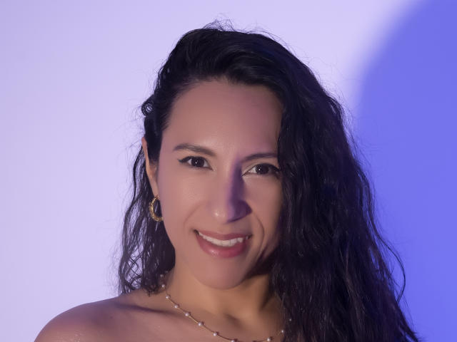 JadeFoster has long, wavy black hair, brown eyes, and a cute smile with shiny teeth. She's wearing gold earrings and a necklace, looking sexy with a soft purple light.