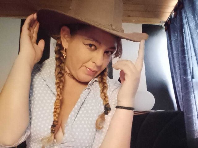 AmandaHotL with braided hair, wearing a tan hat, blue polka-dot shirt, smiling, holding hat, showing her tits slightly, black wristband, earrings, and dark curtains in the background.
