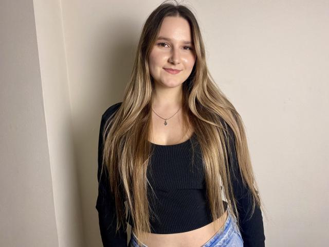 Karpinka with long blonde hair, black crop top, and blue jeans, standing against a plain wall, smiling gently, wearing a small necklace, showing her bare midriff and small tits.