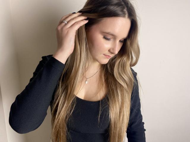 Karpinka with long blonde hair, black top, hand in hair, slight smile, wearing a delicate necklace, looking down, soft light, gentle expression.