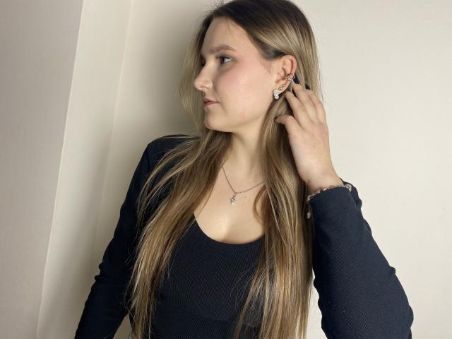 Karpinka with long blonde hair, black top, and earrings, looking sideways, hand touching ear, subtle necklace, soft light, calm expression.