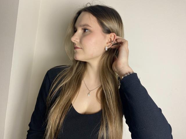 Karpinka with long blonde hair, wearing a black top, touching her ear, looking to the side, showing a necklace and bracelet. Her tits are slightly visible under the top.