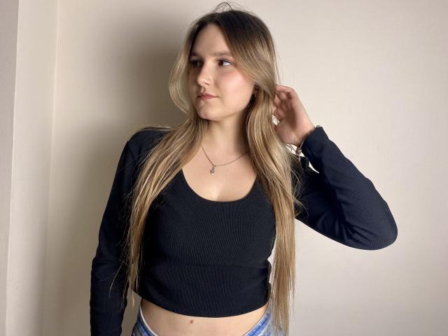 Karpinka with long blonde hair, black crop top, and blue jeans, hand in hair, subtle necklace, standing against a white wall, looking to the side with a slight smile.
