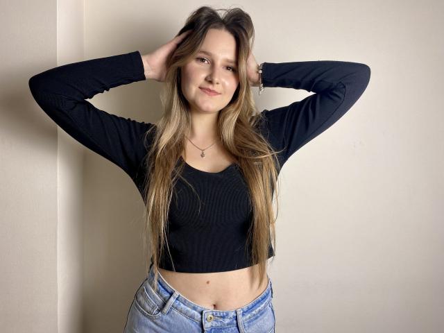 Karpinka with long blonde hair, hands in her hair, black long-sleeve crop top, showing her flat stomach, and blue jeans, smiling slightly.