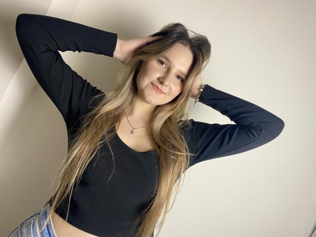 Karpinka with long blonde hair, black long-sleeve crop top, hands in hair, smiling, white wall background, wearing a necklace, light blue jeans, soft lighting.