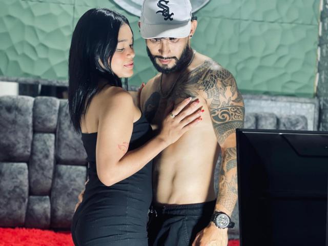 SachaAndFercho, a tattooed guy with a beard, is shirtless, wearing a cap, and black pants, while a woman with black hair and red nails touches his chest.
